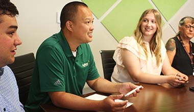 Employee with green shirt and pen talks to interns